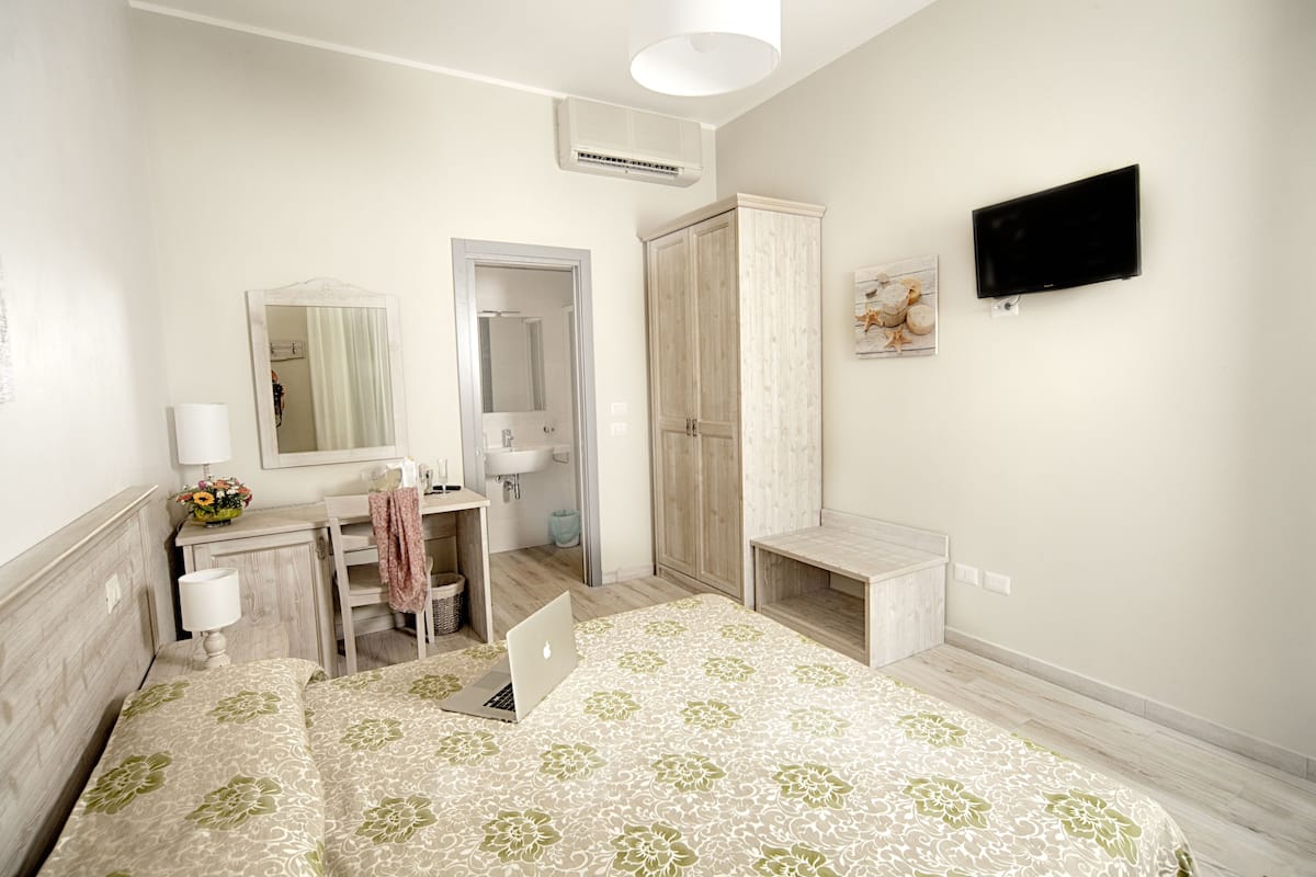 Een hotelkamer met een bed, een bureau met een laptop, een spiegel, een kledingkast, een televisie en een zichtbare badkamer door een open deur.