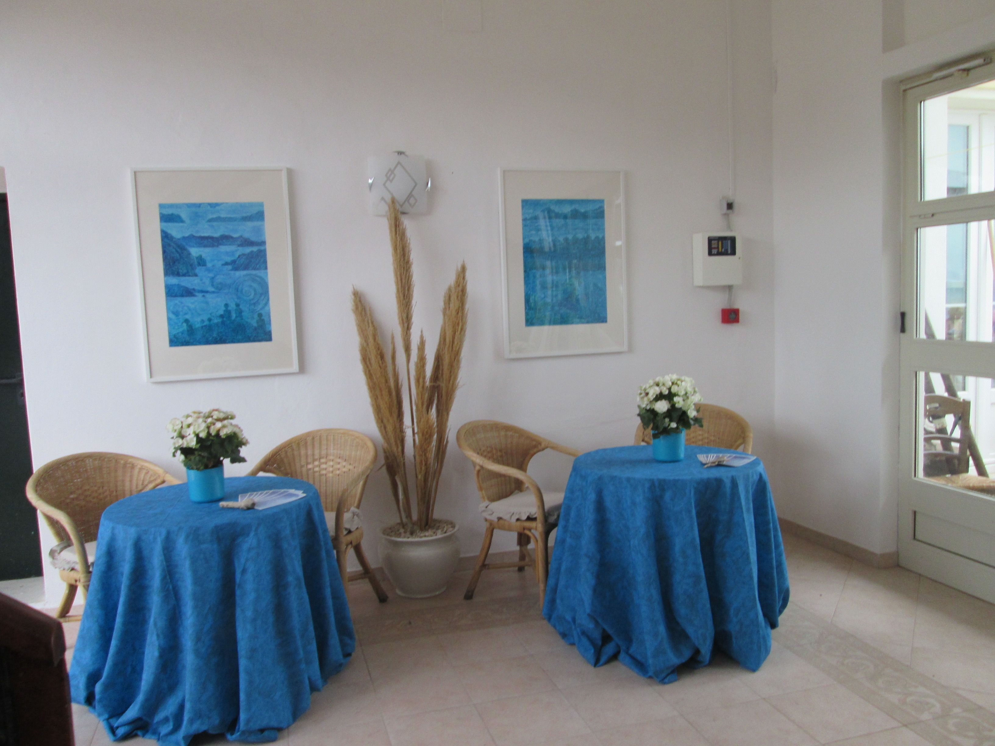 Una habitación con dos mesas cubiertas con manteles azules, cada una con un jarrón de flores blancas. Alrededor de las mesas hay sillas de mimbre y dos cuadros enmarcados en la pared. En el centro hay una planta en maceta con hierbas altas.