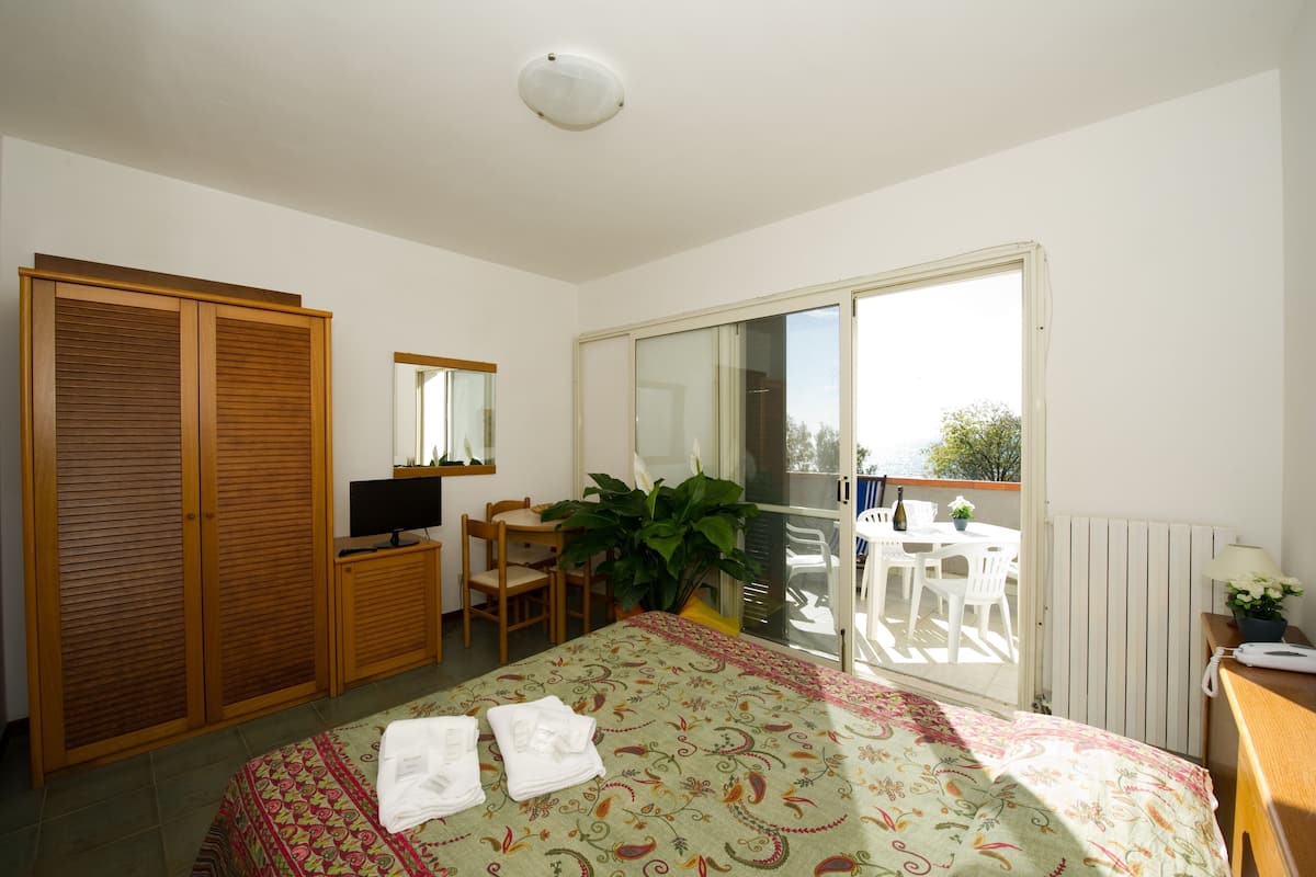 Een slaapkamer met een bed, een bureau, een stoel, een televisie, een kledingkast en een balkon met een tafel en stoelen.