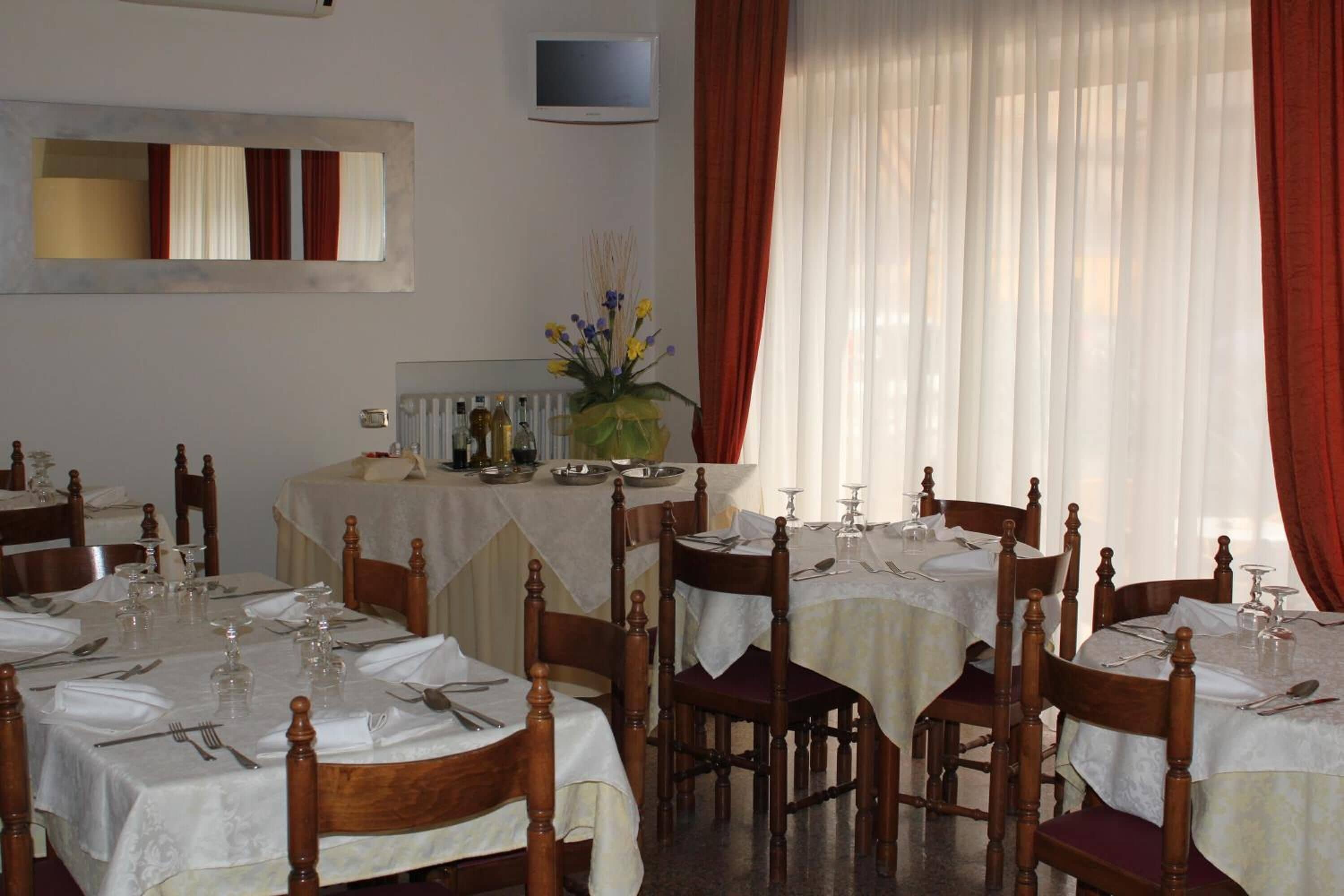 A dining area with multiple tables set for service, wooden chairs, a central buffet table, and a large window with curtains.