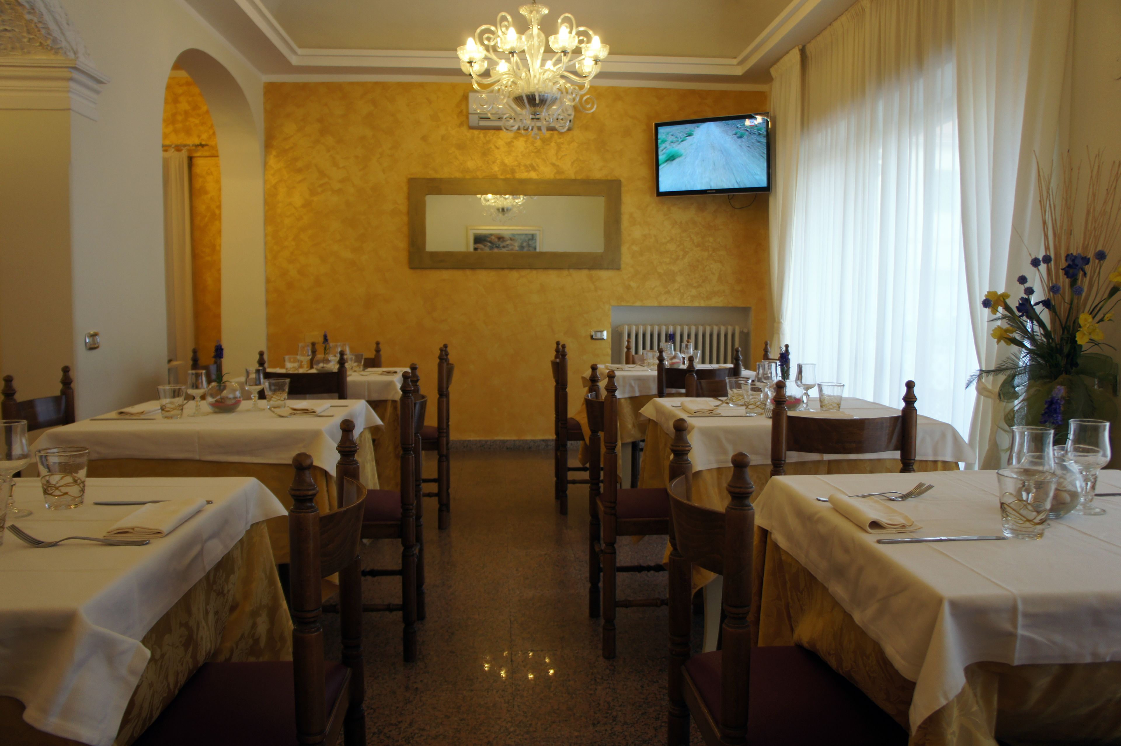Un restaurante con varias mesas, sillas, una araña, un televisor en la pared y un jarrón con flores.