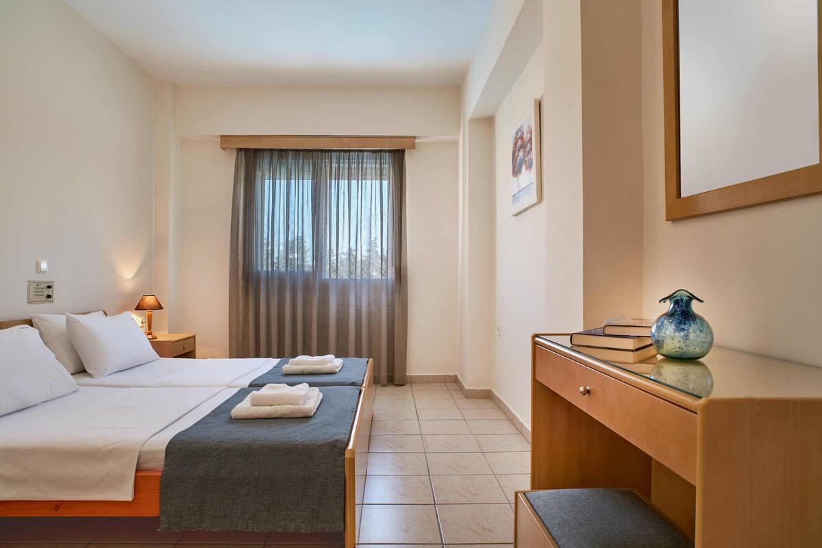 Une chambre d’hôtel avec deux lits, un bureau, une chaise, une lampe et une fenêtre avec des rideaux.