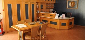 A wooden dining area with a table and chairs, a reception desk, and a wall-mounted shelf with books and a framed picture. - Veronica Hotel (Chania)