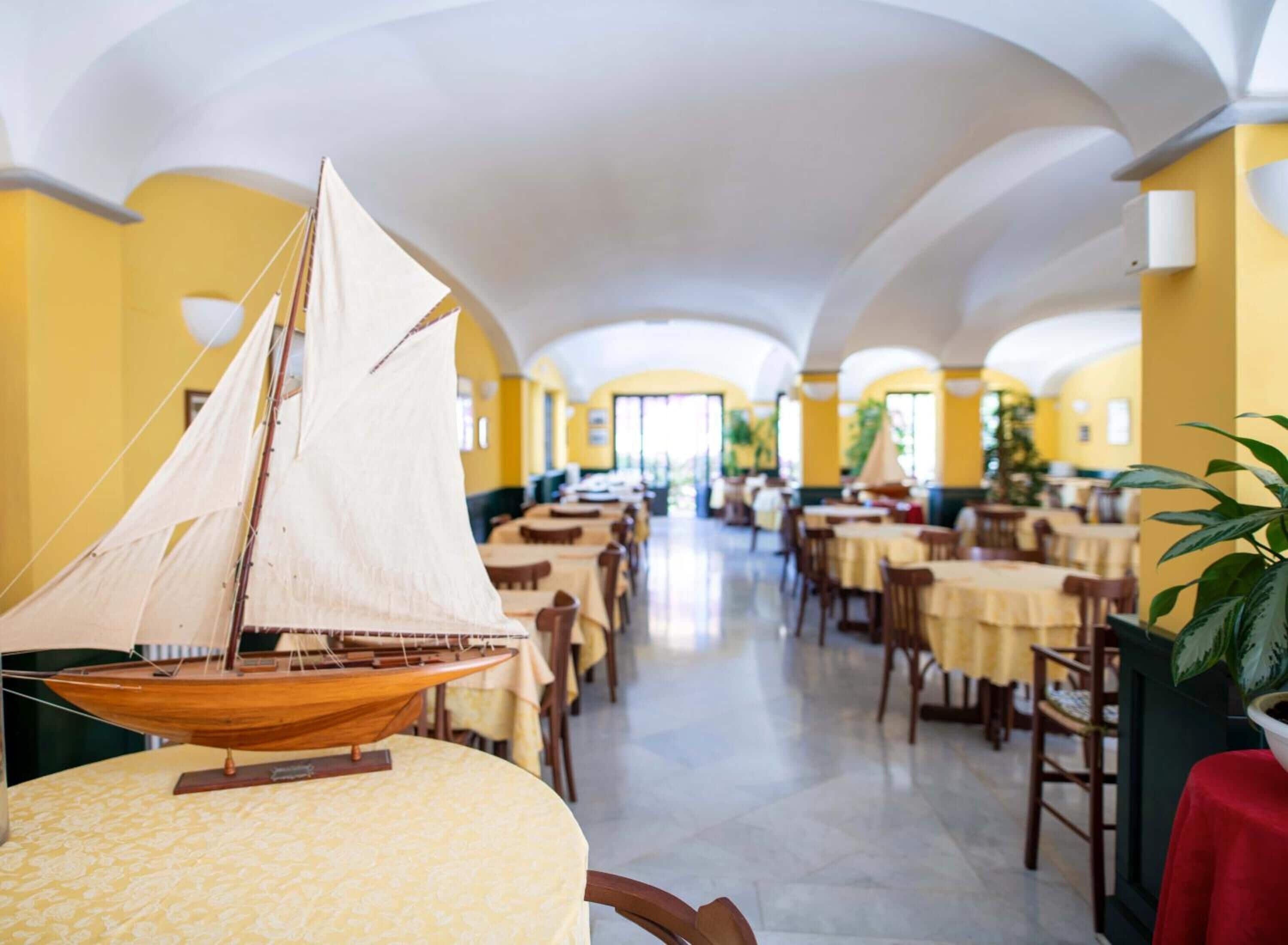 A restaurant with yellow walls and white ceiling arches, featuring tables with yellow tablecloths and a model sailboat on one table.
