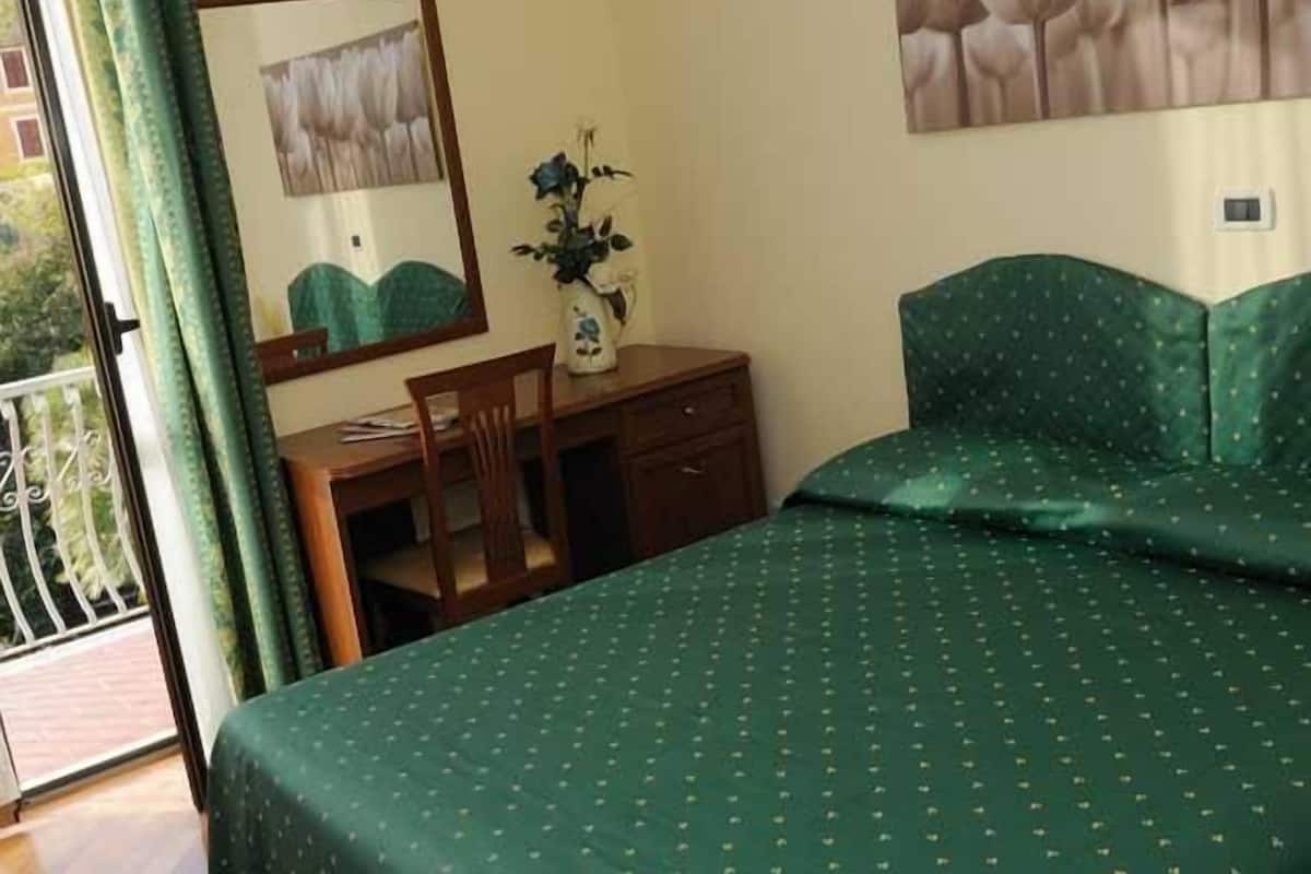 Une chambre à coucher comprenant un lit, un bureau, une chaise, une commode, un vase avec des fleurs et une fenêtre avec des rideaux.