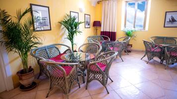 A dining area with wicker chairs and a glass table, surrounded by potted plants and framed pictures on the walls.