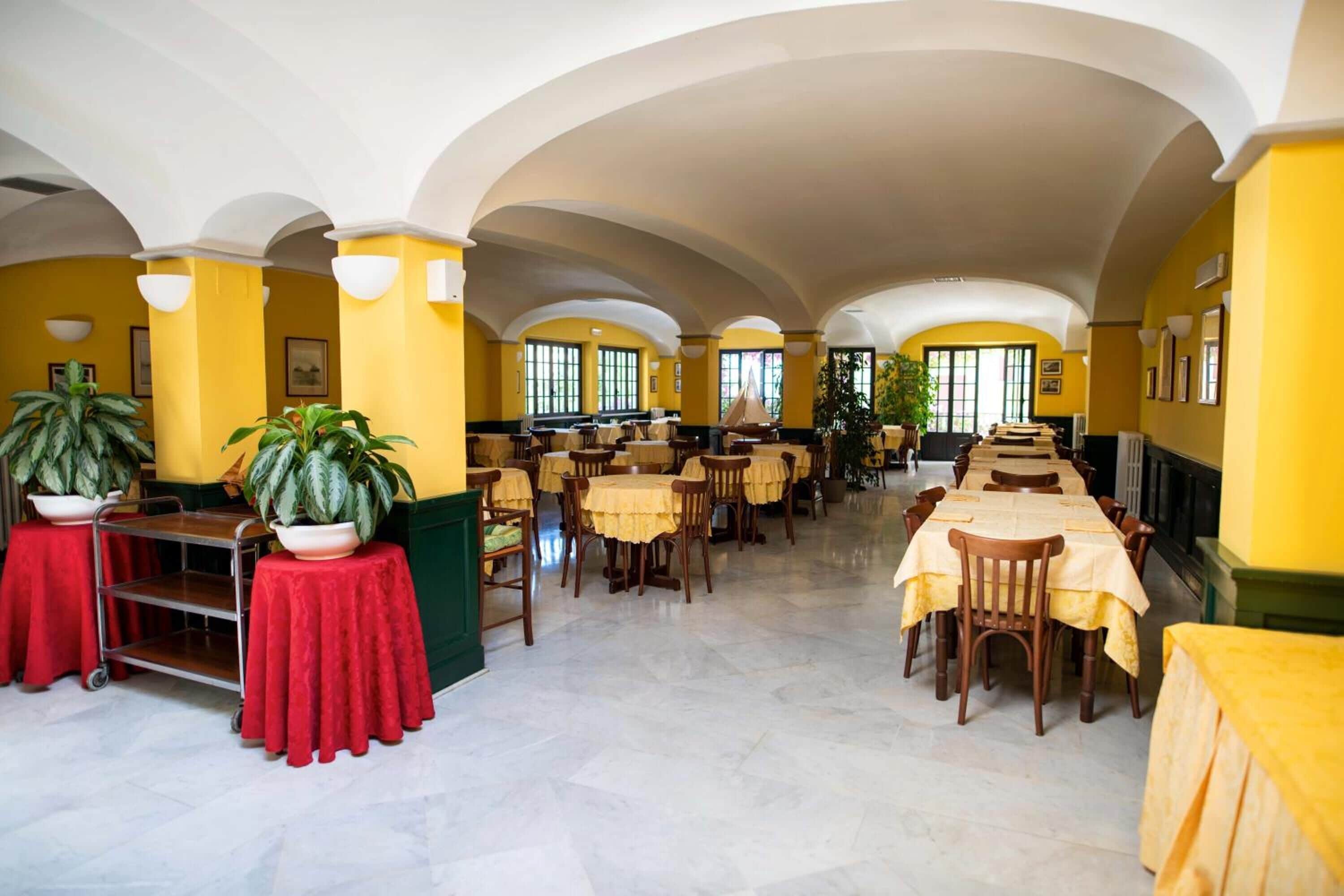 A spacious dining area with high ceilings and yellow walls, featuring multiple tables and chairs arranged for dining.