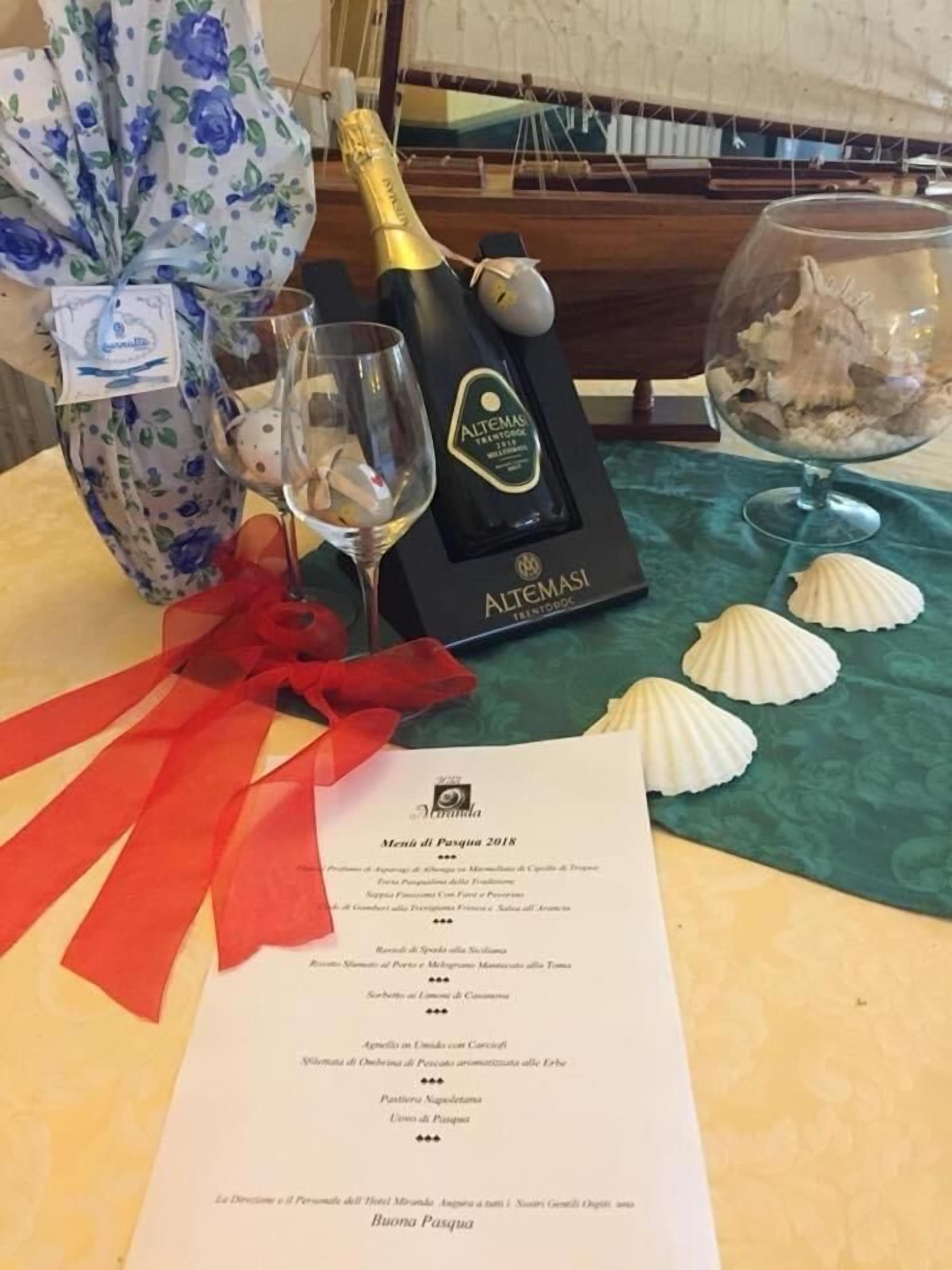 Un menu intitulé « Menu de Pâques 2018 » est posé sur une table recouverte d’une nappe à motifs floraux, avec une bouteille de champagne, deux verres et des coquillages décoratifs.