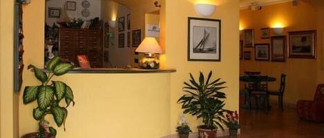 A lobby with a reception desk, potted plants, framed artwork, and a tiled floor.