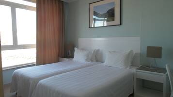Habitación de hotel con dos camas, una mesita de noche, una lámpara, un cuadro enmarcado en la pared y una ventana con cortinas.