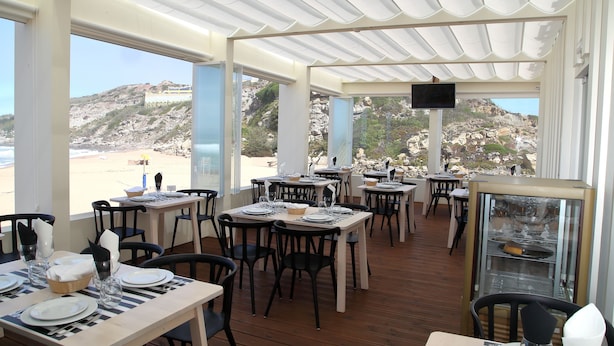En restaurant med borde til spisning, med udsigt over en strand med klippefyldt terræn.