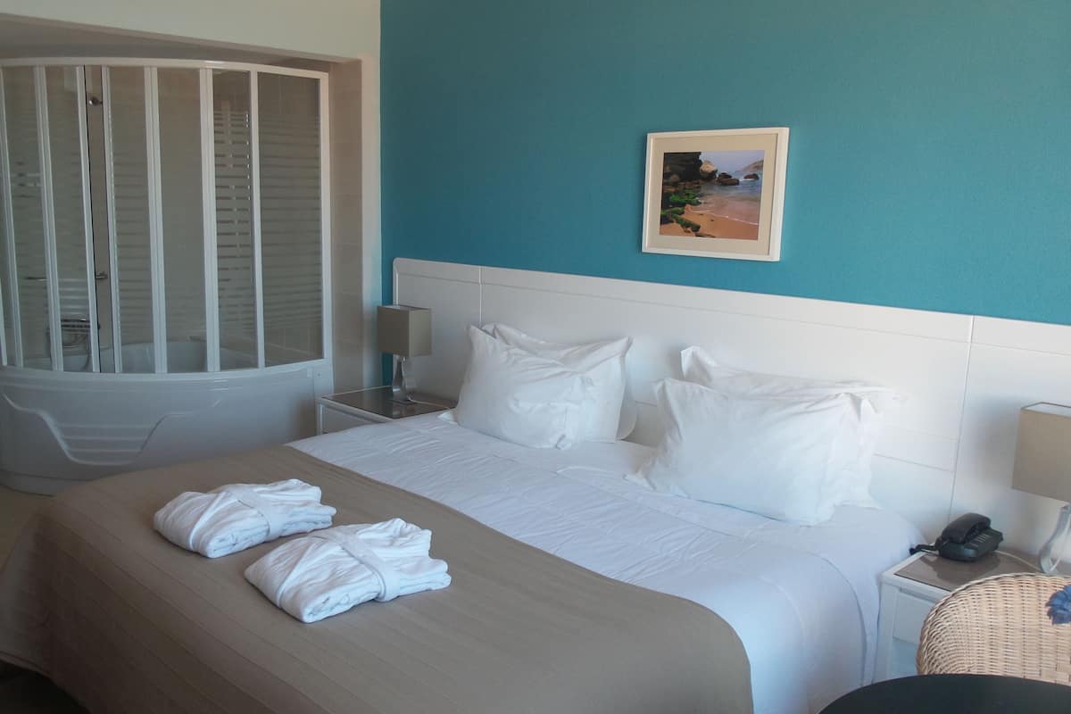 A hotel room with a bed, a bathroom with a shower, a picture on the wall, and a phone on the nightstand.