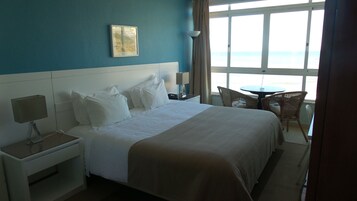 Habitación de hotel con una cama grande, una mesita de noche con lámpara, una silla y vista al mar por la ventana.