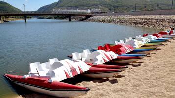 Uma fileira de coloridos barcos a pedalinho alinhados em uma praia arenosa, ao lado de um rio calmo, com uma ponte e colinas ao fundo.
