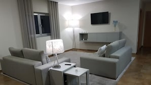 Una sala de estar moderna con dos sofás grises, una mesita blanca, una lámpara de pie y un televisor montado en la pared.