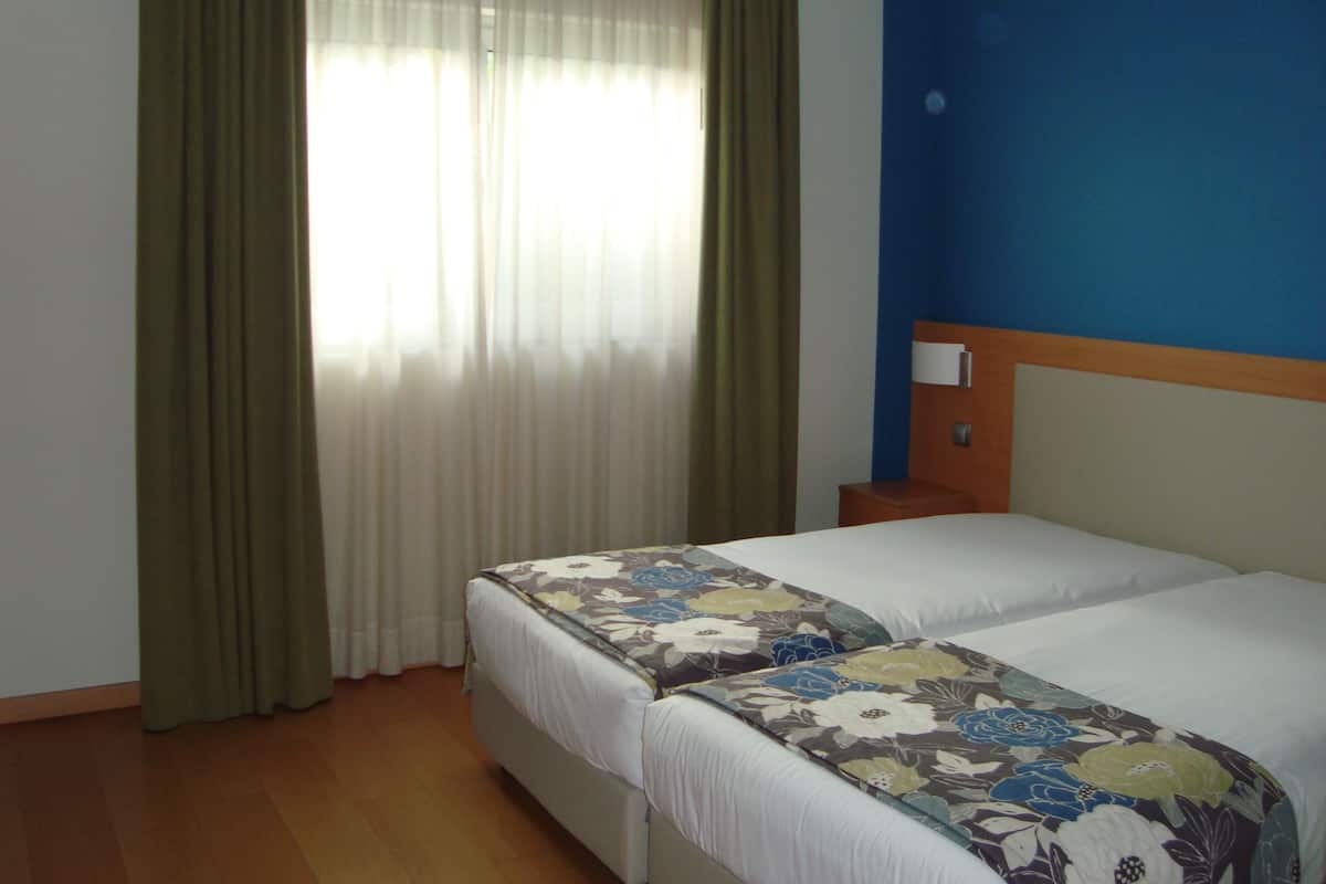 A hotel room with two beds, a wooden headboard, a nightstand, and a window with sheer curtains.
