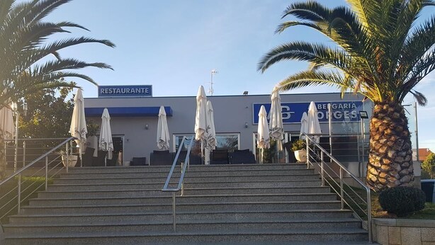 Un edificio con un letrero que dice "RESTAURANTE" y "BERGARIA GES", con mesas al aire libre y palmeras.