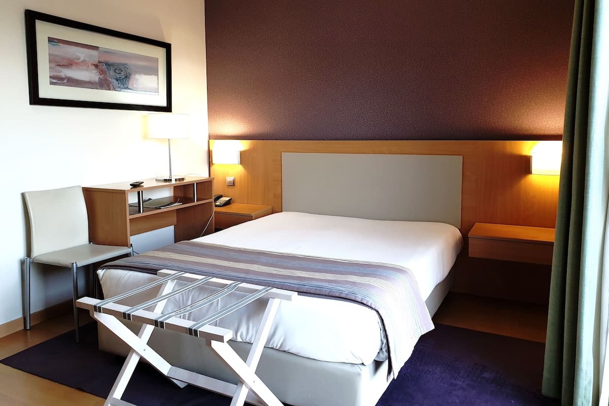 A hotel room with a bed, bedside tables, a desk, a chair, a lamp, a telephone, and a picture on the wall.