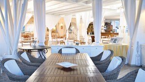 Un área de comedor al aire libre con mobiliario de mimbre y cortinas blancas, con vista al interior de un restaurante.