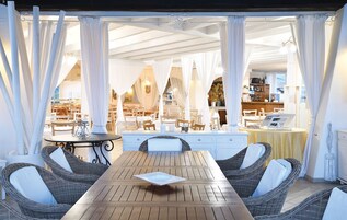 Un área de comedor al aire libre con mobiliario de mimbre y cortinas blancas, con vista al interior de un restaurante.