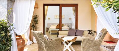 A cozy outdoor seating area with wicker furniture, a wooden table, and a view of a bedroom with a bed and a window.