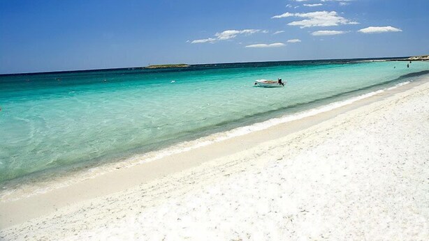 Una playa con agua turquesa cristalina, una orilla arenosa y un barco a lo lejos.