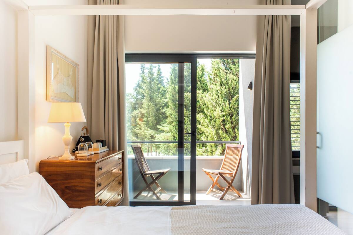 Ein Hotelzimmer mit Bett, einem Holzschrank, einer Lampe, einem gerahmten Bild an der Wand und einem Balkon mit zwei Stühlen und Blick auf Bäume.