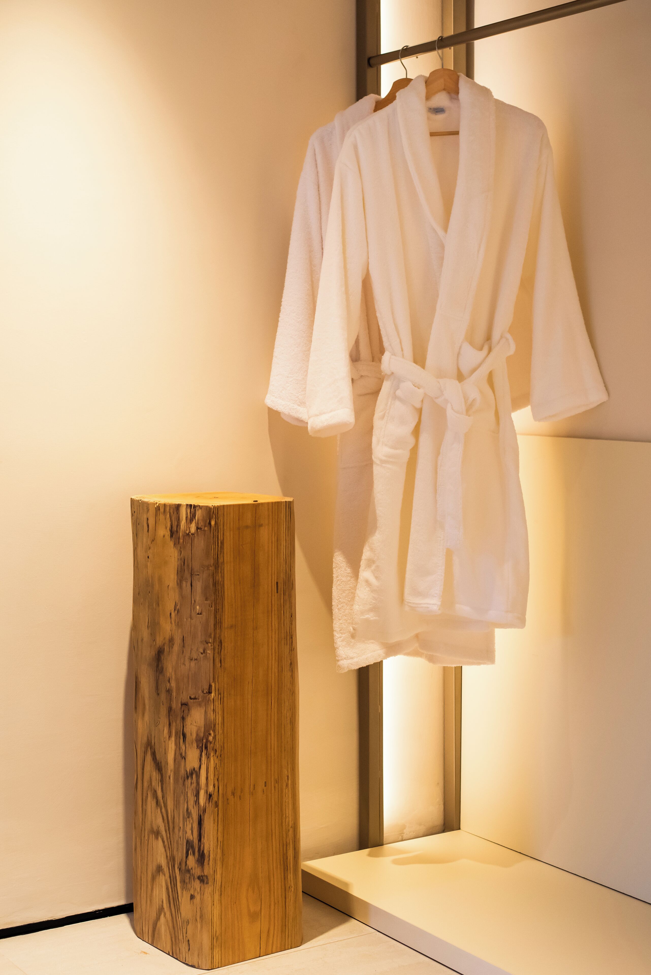 A white bathrobe hanging on a wooden hanger next to a wooden stool in a well-lit room.