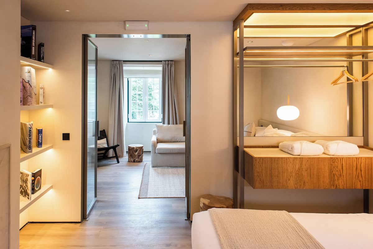 Ein modernes Hotelzimmer mit einem Holznähtischchen, einem großen Spiegel und einem Bett mit weißer Decke. Außerdem ist ein kleiner Holzschemel und ein Fenster mit Vorhängen vorhanden.