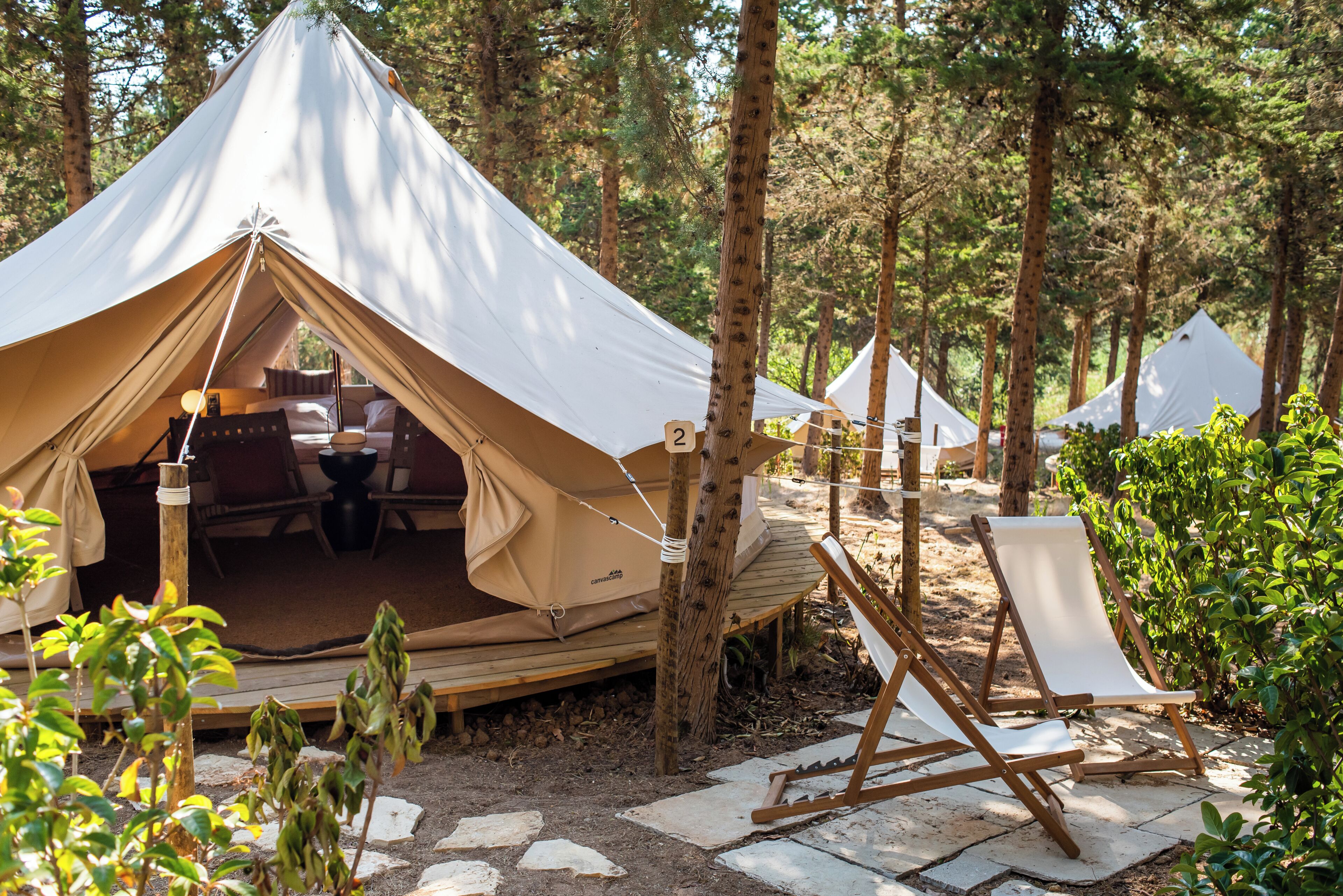 En campingplats i ett skogsområde med ett stort tält, en solstol och en stig av sten.