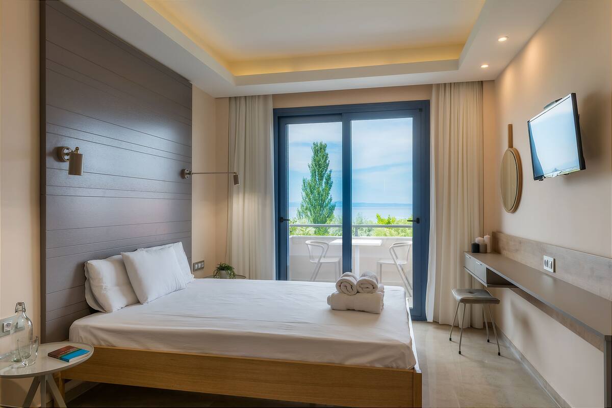 A hotel room with a bed, a desk, a TV, and a balcony with chairs.