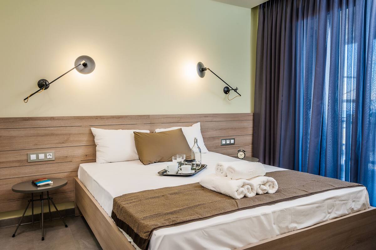 A neatly made bed with a brown blanket, a tray with a bottle, glasses, and a cup, and a small table with a book beside it.