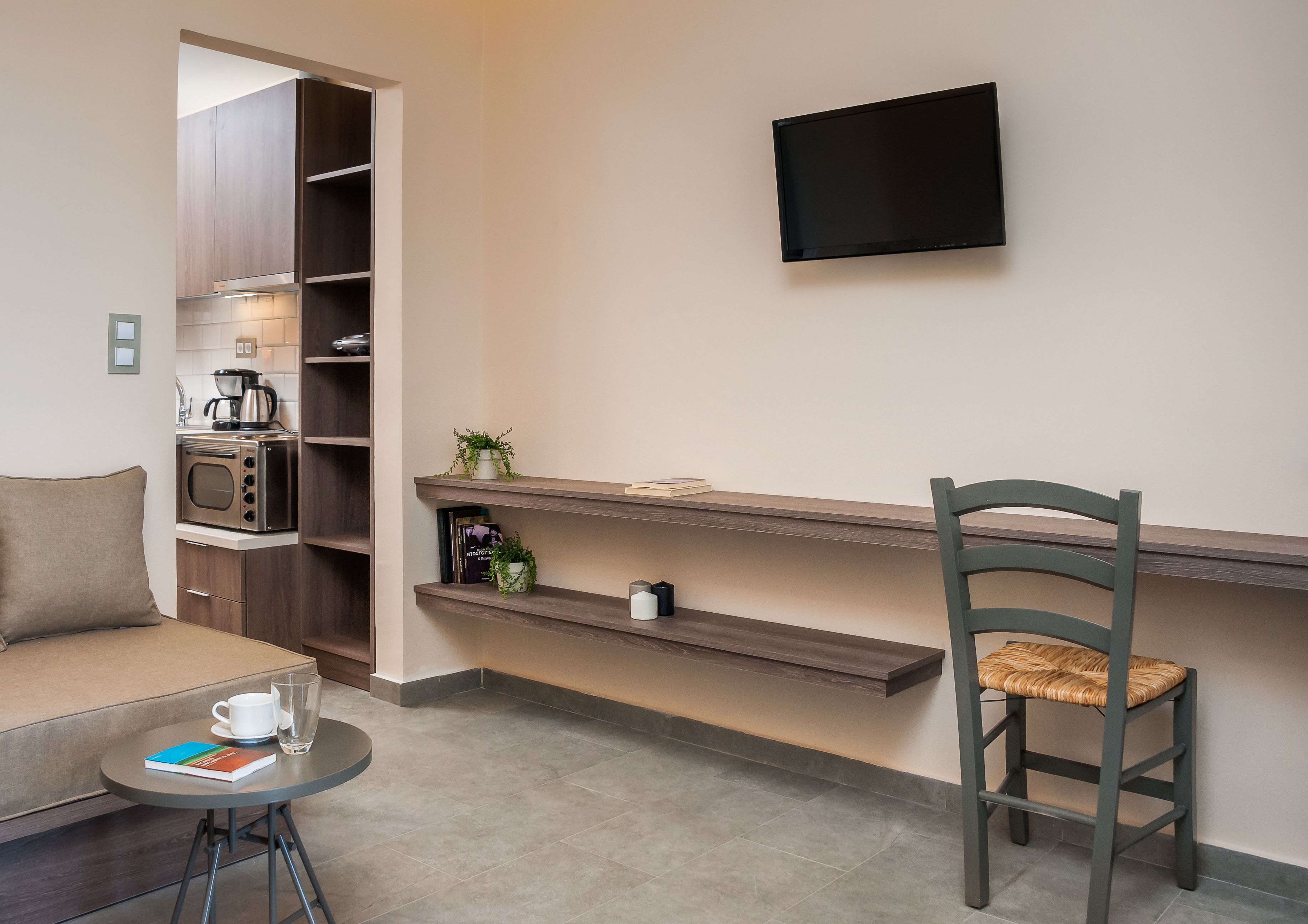 A modern kitchenette with a microwave, toaster, and coffee maker. There is a TV mounted on the wall, a chair with a woven seat, and a small table with a book and a cup.