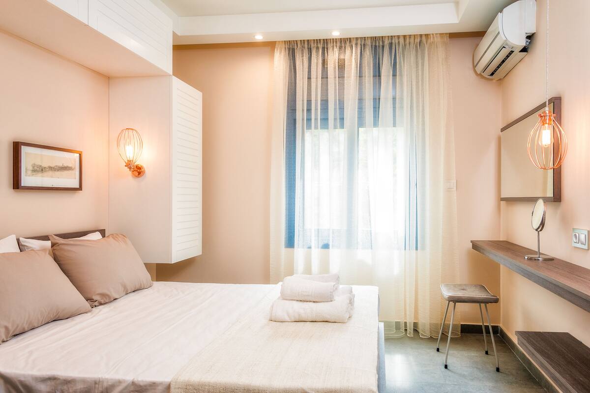 A bedroom with a bed, a bedside table, a mirror, and a window with sheer curtains.