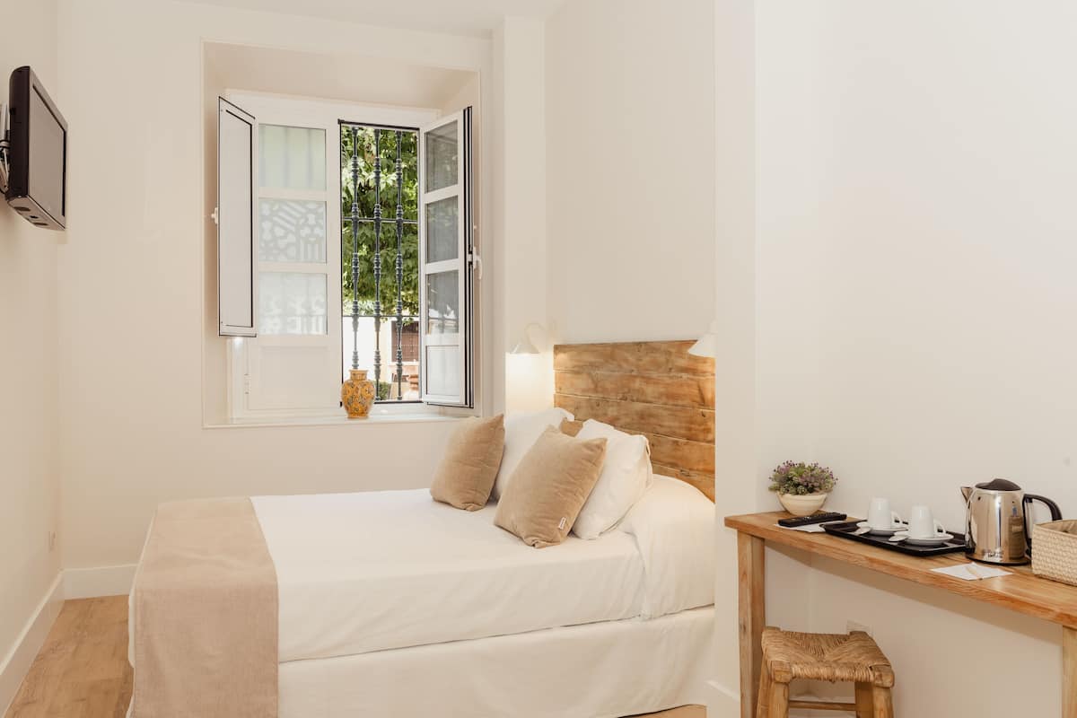 A bedroom with a bed, a wooden headboard, a bedside table with a lamp, a window with a view of greenery, and a small stool.