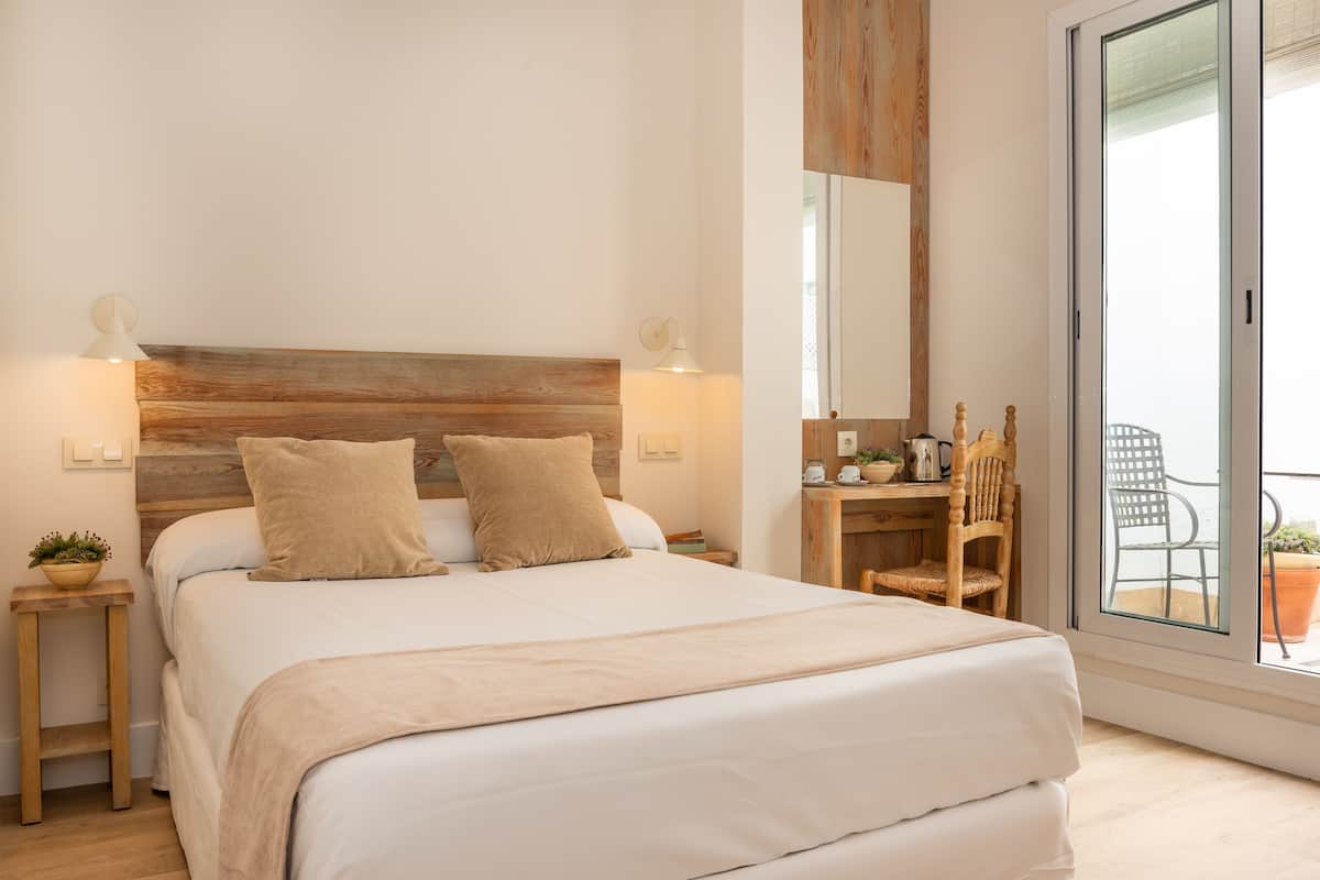 A bedroom with a wooden headboard, two bedside lamps, a bed with white linens, a small table with a plant, and a view of the outside balcony through a glass door.