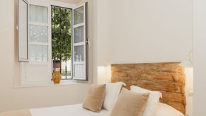 A bedroom with a wooden headboard, a bed with white and beige bedding, a window with a view of greenery, and a vase on the windowsill. - Hotel Boutique Elvira Plaza (Seville)