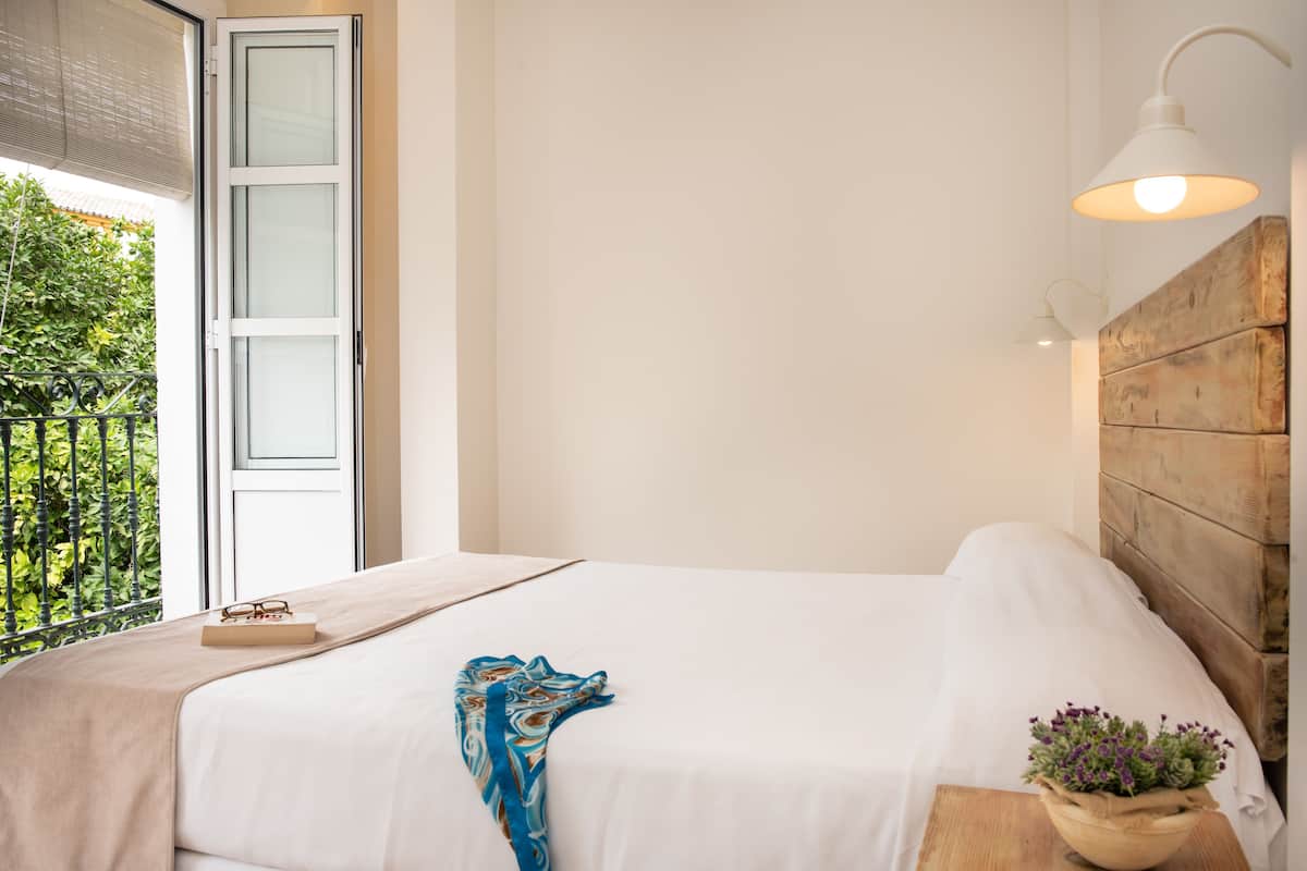 A bedroom with a bed, a bedside table with a plant, a window with a view of greenery, and a door to an outdoor area.