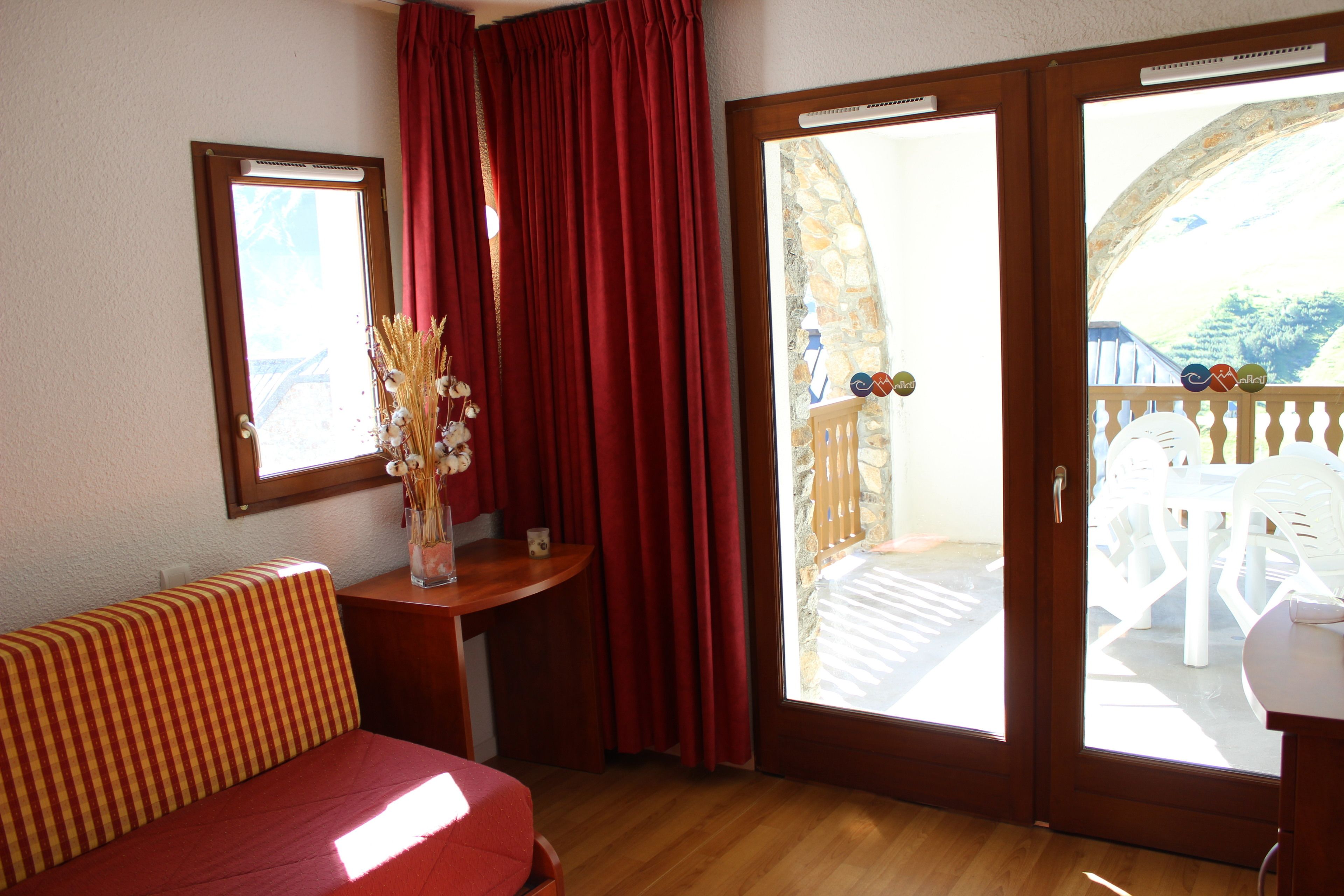 Una habitación con suelo de madera, un sofá a rayas, una mesita con un jarrón de flores secas y un ventanal corredero que da a un área exterior con una mesa y sillas blancas.