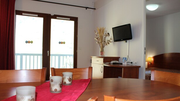 Un comedor con mesa de madera, mantel rojo y dos velas decorativas. Se observa un televisor montado en la pared y un pequeño escritorio con lámpara.