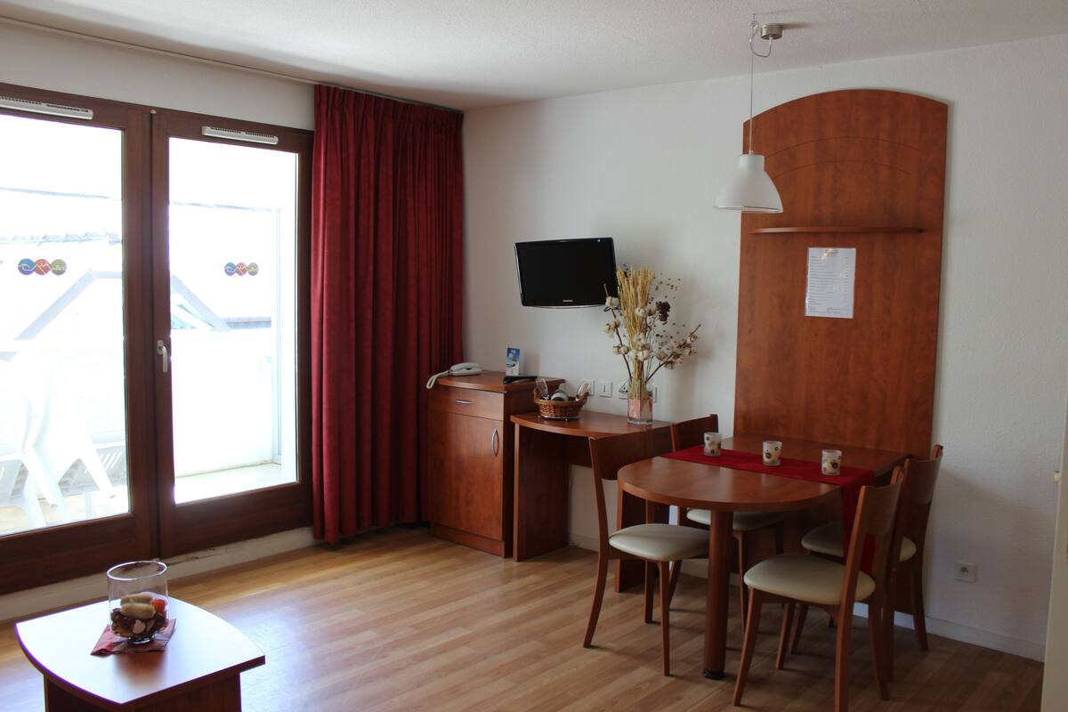 A room with a wooden desk, a dining table with chairs, a television mounted on the wall, and a window with red curtains.