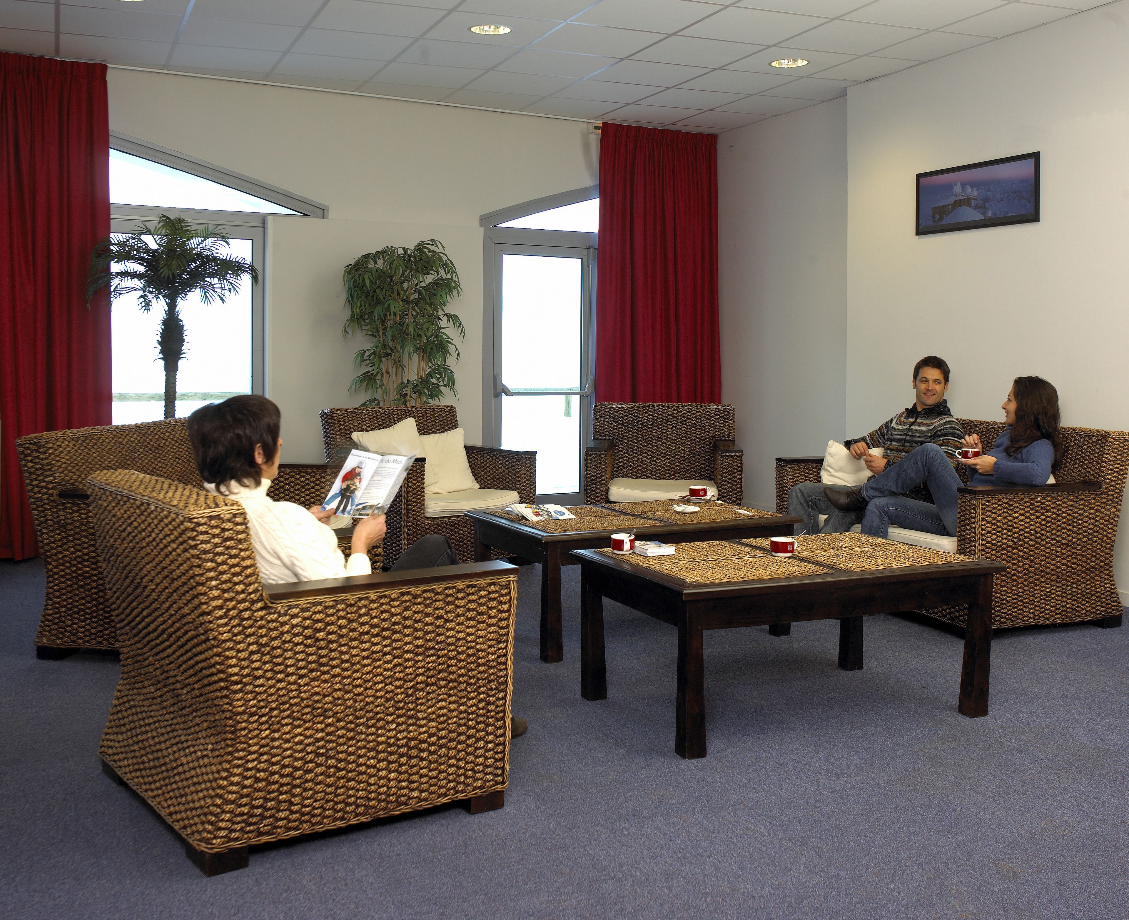 A room with wicker furniture, a coffee table, and a large window with red curtains.