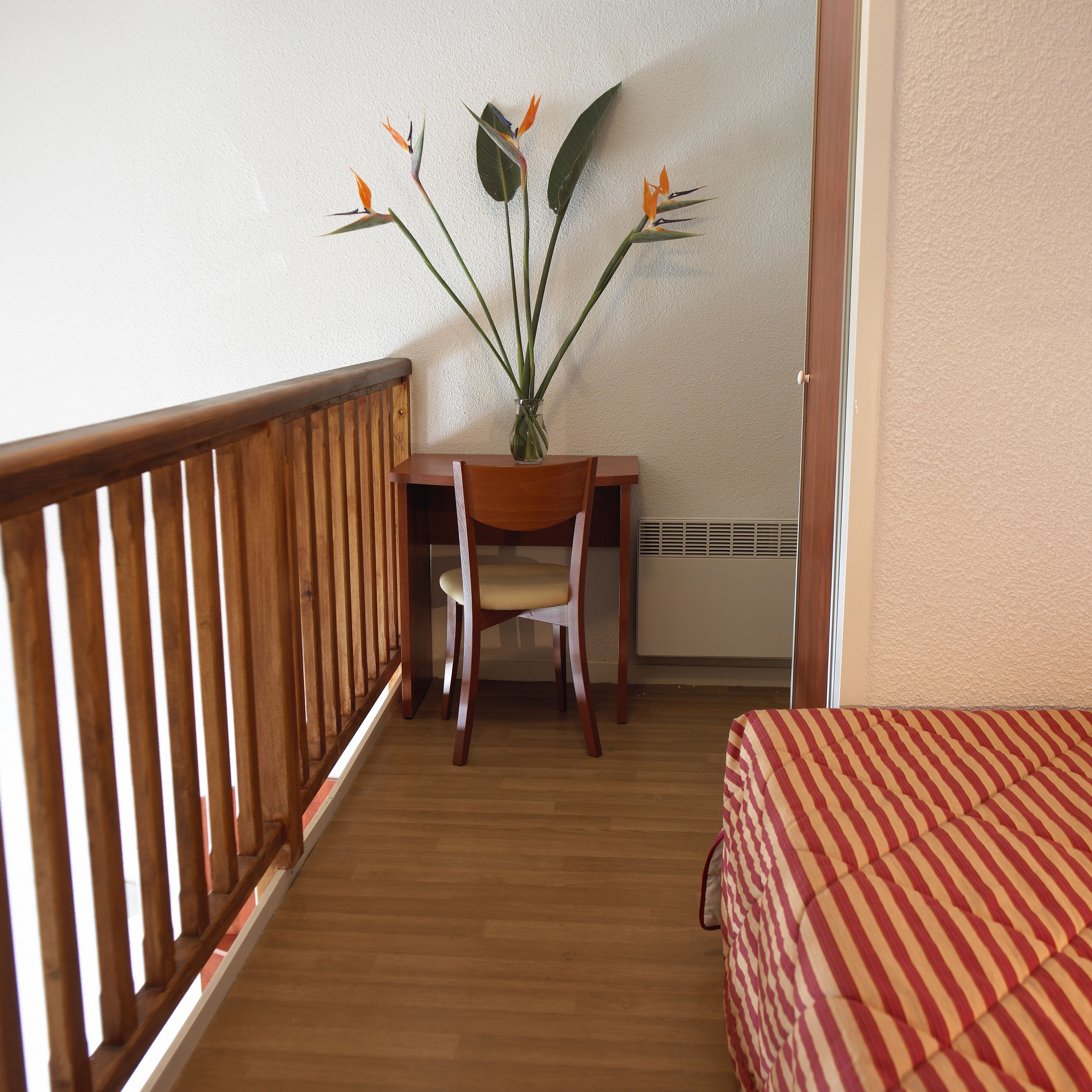 A room with a wooden desk, a chair, a striped bed, and a plant with orange flowers.