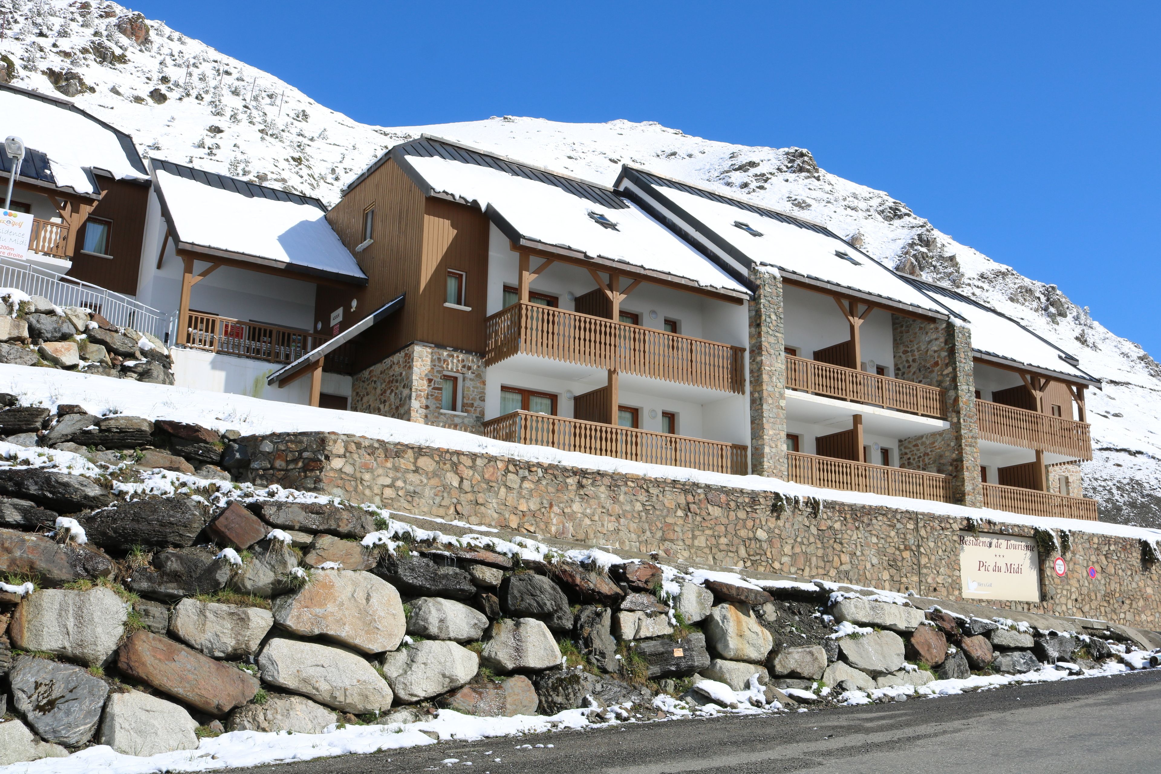 A multi-story hotel with stone walls and wooden balconies, set against a snowy mountain backdrop.