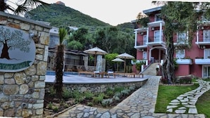 A stone wall with a tree logo and a pathway leading to a building with balconies and a garden. - Vassilis Guest House (Parga)