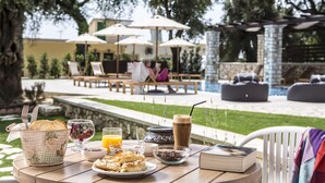 A breakfast table with croissants, coffee, and juice, set outdoors with a pool and lounge chairs in the background. - Vassilis Guest House (Parga)