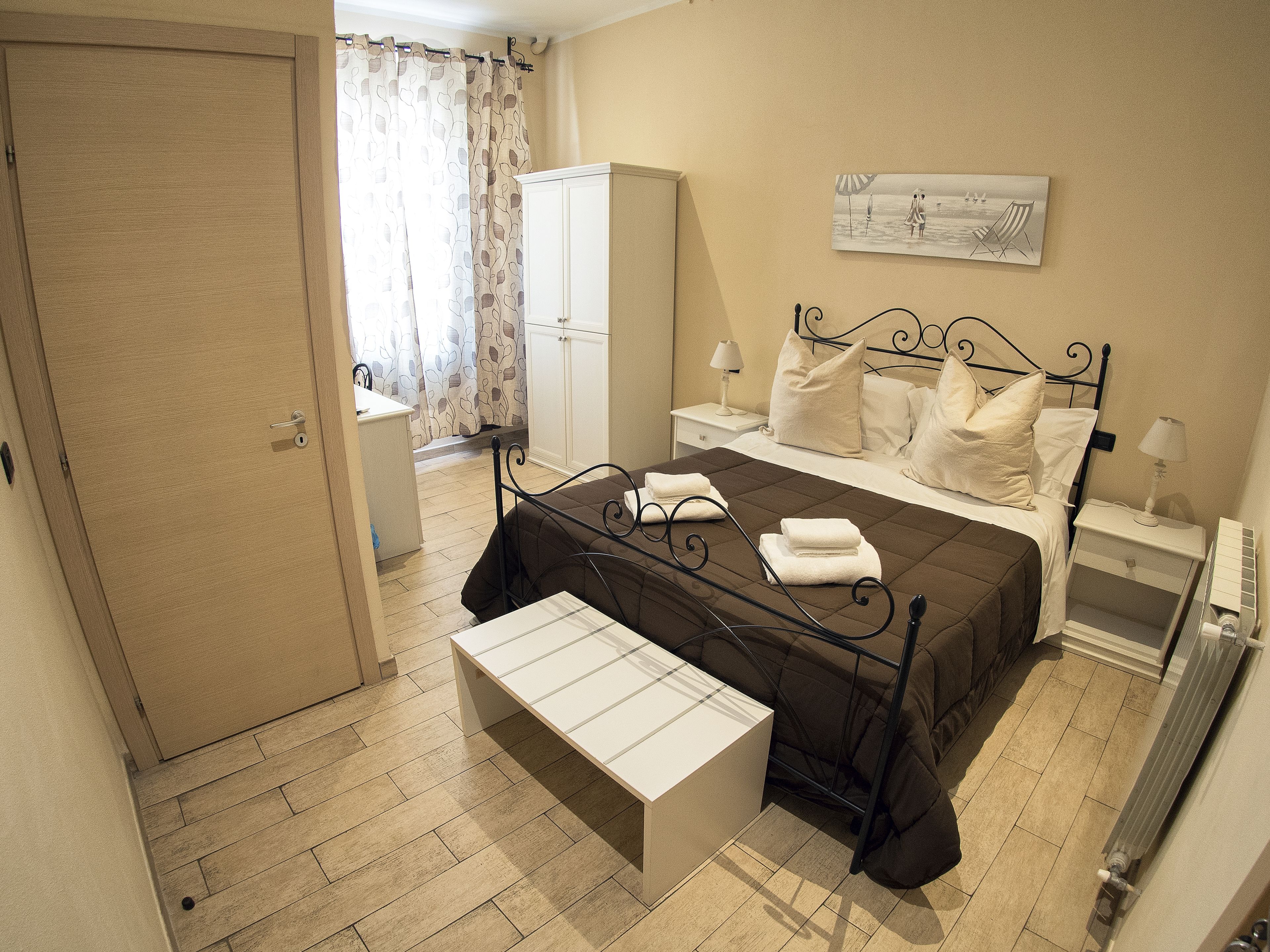 A hotel room with a bed, bedside tables, a wardrobe, a bench, and a painting on the wall.
