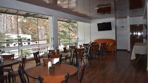A restaurant with round tables and chairs, large windows with a view of trees and buildings, and a flat-screen TV mounted on the wall.