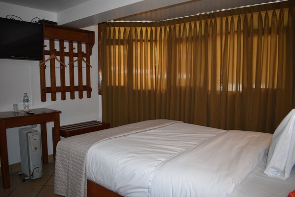 Una habitación de hotel con una cama, un televisor montado en la pared, un escritorio de madera y un radiador.