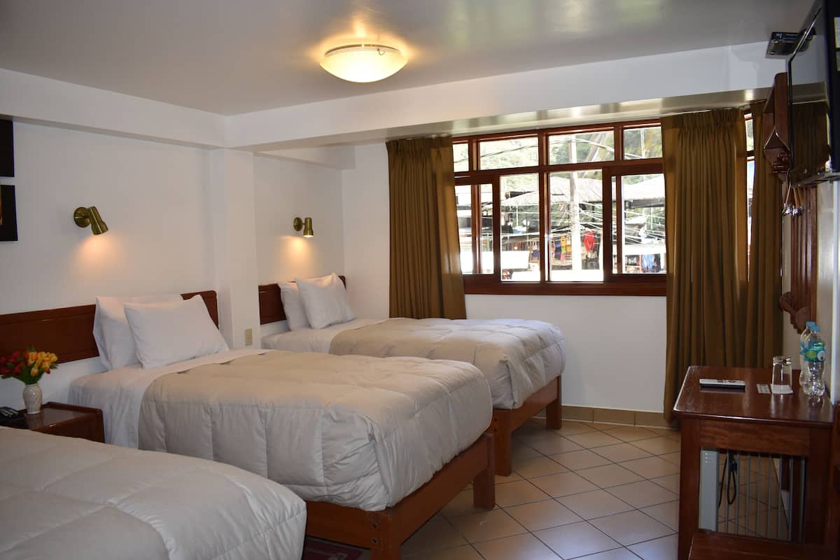 A hotel room with two beds, a wooden nightstand with a vase of flowers, a wooden table with a water bottle, and a window with curtains.